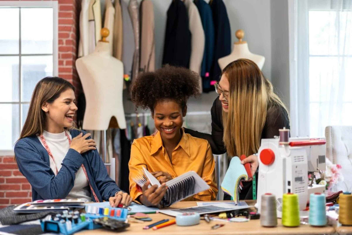 Three students in a sewing or design studio with fabric, mannequins, and sewing tools.