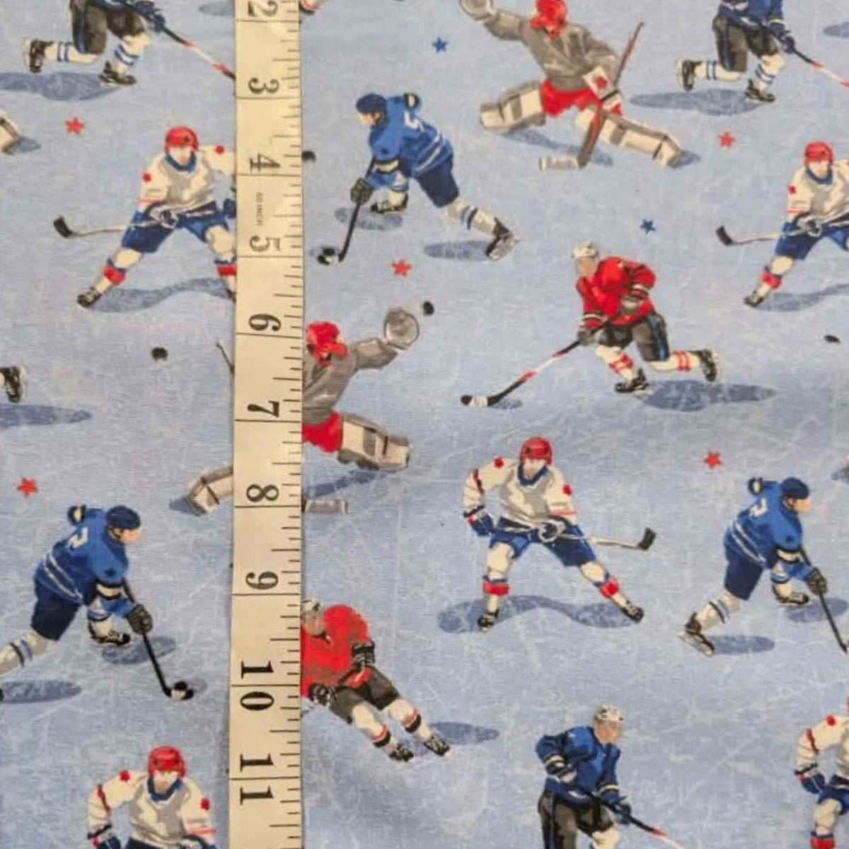 A close-up of patterned fabric featuring illustrated ice hockey players on a light blue ice background, with a vertical ruler overlaid to show scale.