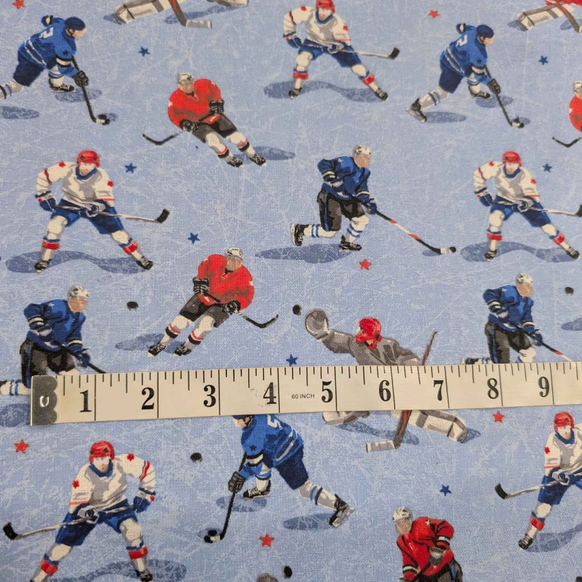 A close-up of patterned fabric featuring repeated ice hockey players in blue, red, white, and gray on a light blue background, with a measuring tape laid across the lower portion to show scale.