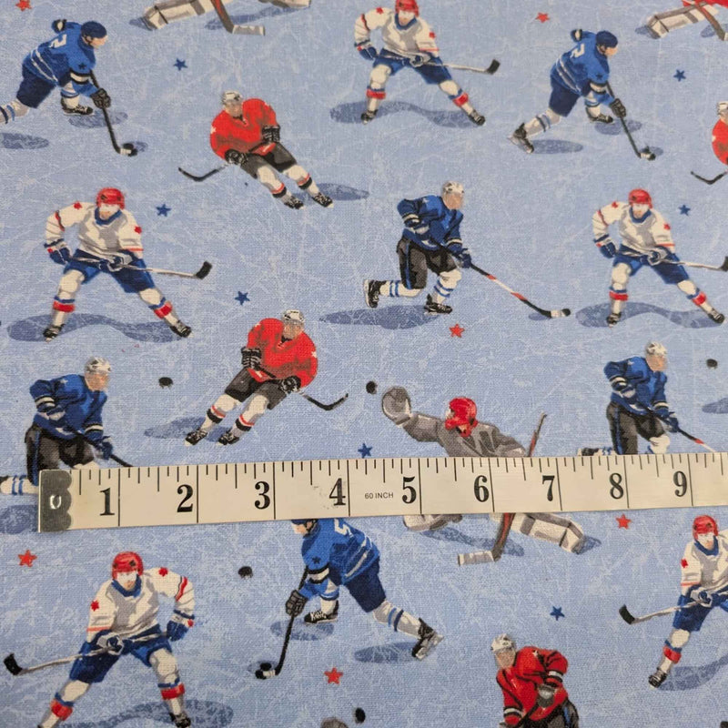A close-up of patterned fabric featuring repeated ice hockey players in blue, red, white, and gray on a light blue background, with a measuring tape laid across the lower portion to show scale.