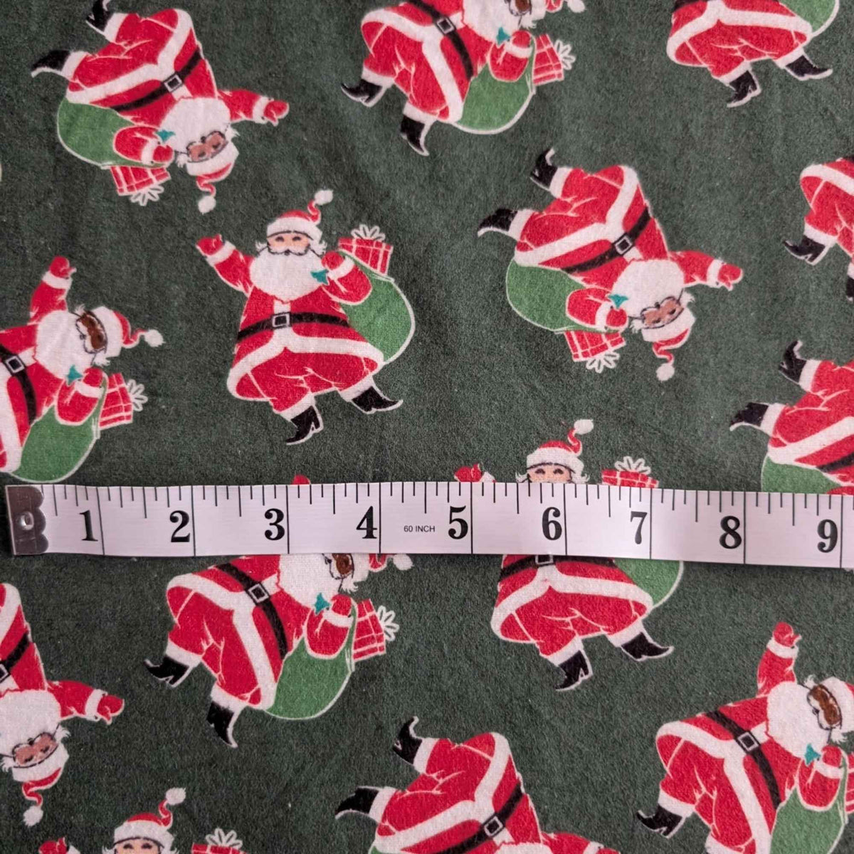 Dark green cotton flannel fabric with tossed Santa Claus figures in red suits carrying green sacks with wrapped presents.