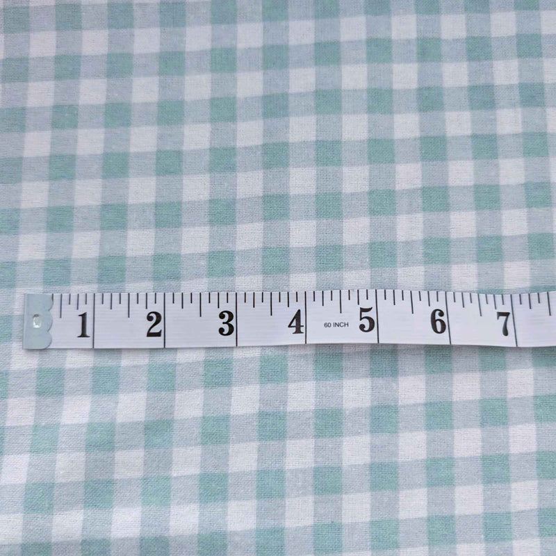 A close-up of a light blue and white gingham fabric with a white measuring tape ruler laid horizontally across the center for scale.